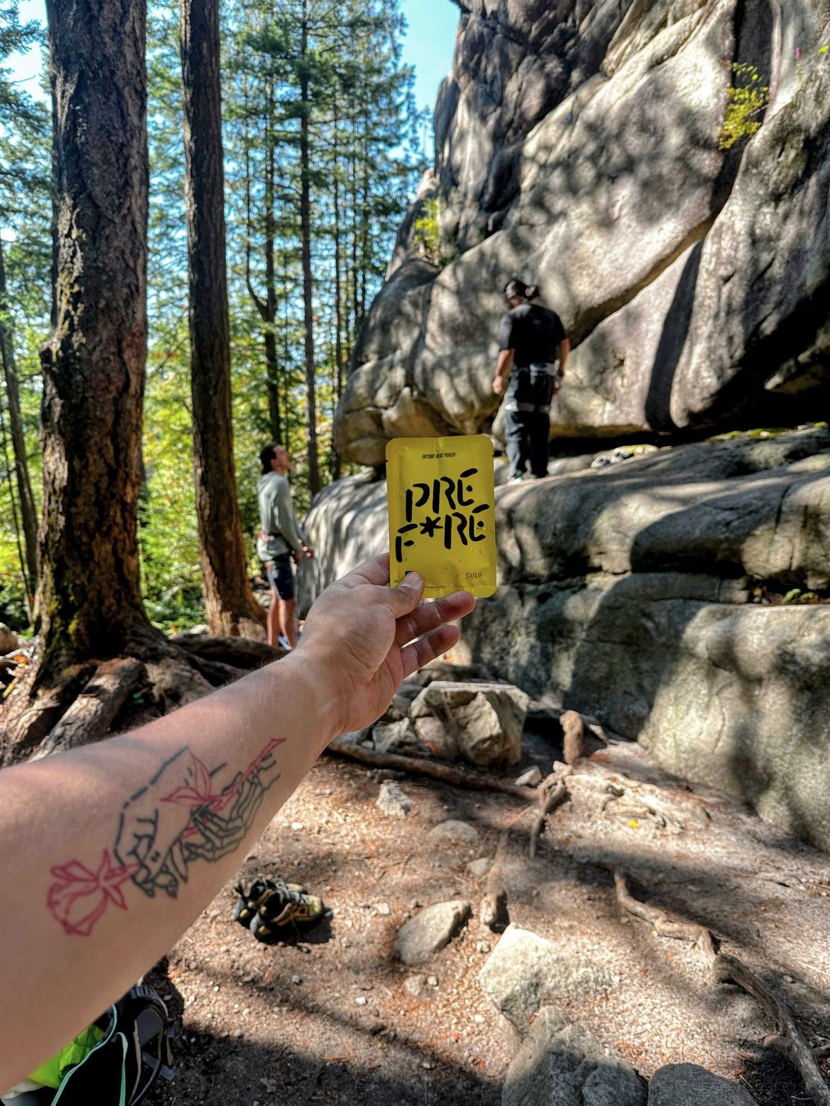 Tattooed arm holding a yellow PREFIRE pouch at the crag with climbers behind